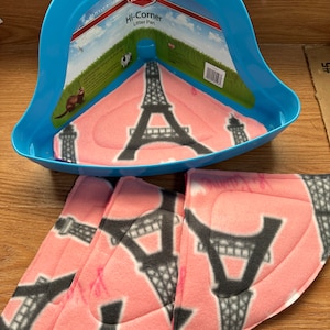 May include: A blue Kaytee Hi-Corner litter pan with a pink Eiffel Tower design. The pan includes matching pink pads with the same Eiffel Tower pattern. The packaging shows illustrations of small animals.