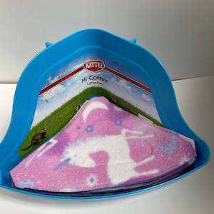 May include: Blue plastic corner litter pan with a pink and white fleece liner with a unicorn and moon design. The pan has a label that reads "Kaytee Hi-Corner Litter Pan".