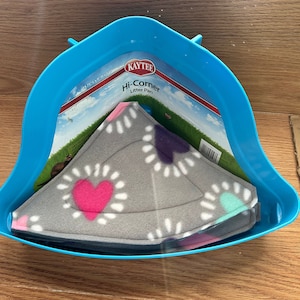 May include: A blue plastic Kaytee Hi-Corner litter pan with a gray fleece corner bed. The bed has a floral pattern with pink, purple, and teal accents. The packaging is visible inside the pan.