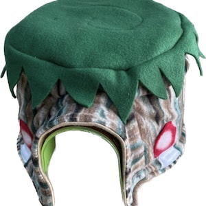 May include: A green and brown felt hat shaped like a tree stump. The hat has a green felt top with a scalloped edge and a brown felt base with a camouflage pattern. There are two red and white circles on the side of the hat.