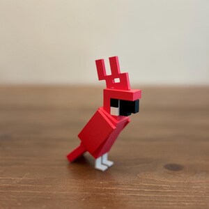 May include: A red, geometric bird figurine with a black beak, white feet, and a distinctive crest. The bird is constructed from blocky shapes, giving it a pixelated appearance. The figurine is set against a neutral background.