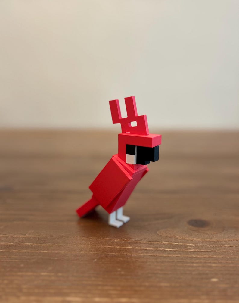 May include: A red, geometric bird figurine with a black beak, white feet, and a distinctive crest. The bird is constructed from blocky shapes, giving it a pixelated appearance. The figurine is set against a neutral background.