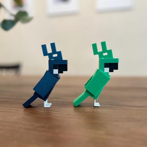 May include: Two geometric parrot figurines. One is dark blue, the other is green. Both have black beaks and white feet. They are positioned on a wooden surface, with a blurred background.