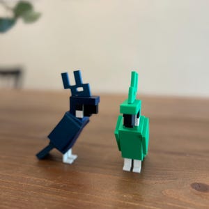 May include: Two blocky bird figurines. One is dark blue with a black head and white accents. The other is green with a black head and white accents. Both are standing on a wooden surface.