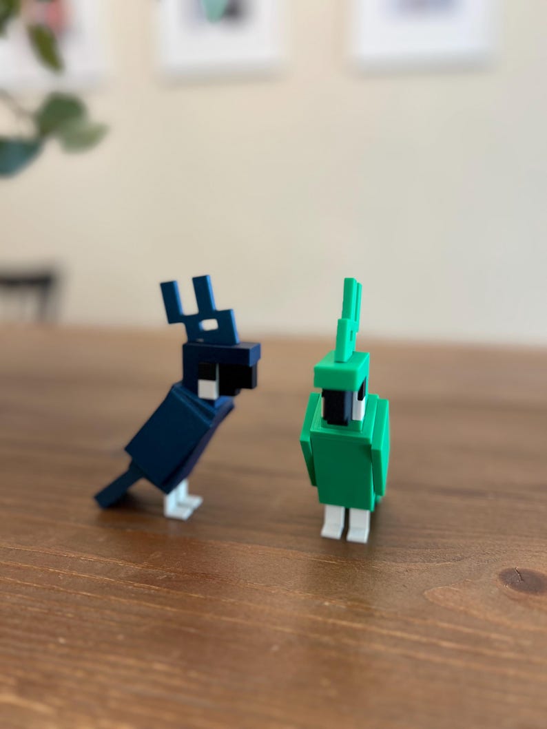 May include: Two blocky bird figurines. One is dark blue with a black head and white accents. The other is green with a black head and white accents. Both are standing on a wooden surface.