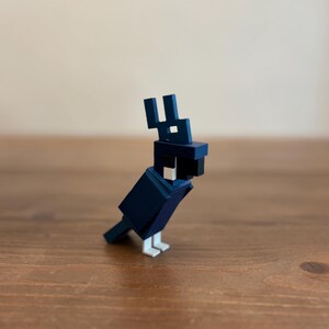May include: A geometric, navy blue bird figurine with white feet and a black beak. The bird has a blocky, pixelated design, reminiscent of a video game character. The figurine is made of a solid material and sits on a wooden surface.