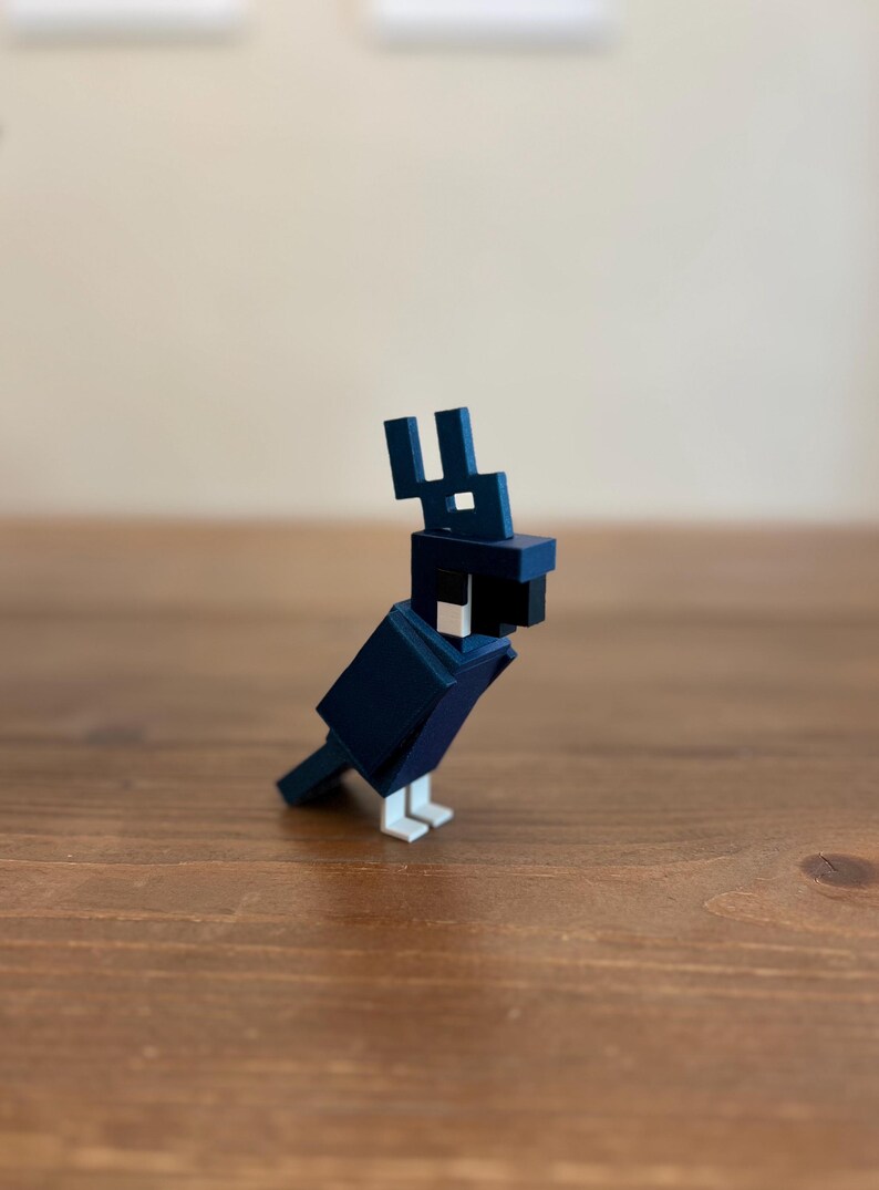 May include: A geometric, navy blue bird figurine with white feet and a black beak. The bird has a blocky, pixelated design, reminiscent of a video game character. The figurine is made of a solid material and sits on a wooden surface.