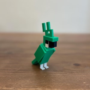 May include: A green, blocky parrot figurine with a black beak and white feet. The parrot has a distinctive crest and is made of geometric shapes. The figurine is displayed on a wooden surface.