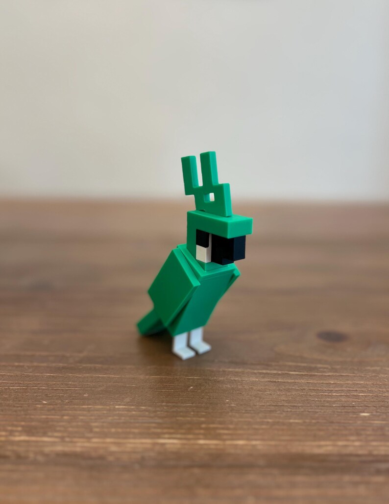 May include: A green, blocky parrot figurine with a black beak and white feet. The parrot has a distinctive crest and is made of geometric shapes. The figurine is displayed on a wooden surface.