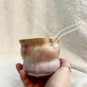 Puede incluir: Una pipa de cerámica hecha a mano con una pajita de cristal transparente. La pipa es una mezcla de colores rosa, morado y beige. La pipa se sostiene en una mano.