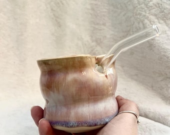 Puede incluir: Una pipa de cerámica hecha a mano con una pajita de cristal transparente. La pipa es una mezcla de colores rosa, morado y beige. La pipa se sostiene en una mano.