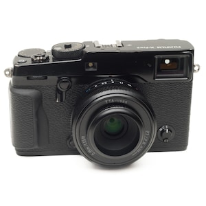 May include: A black Fujifilm X-Pro3 digital camera with a TTArtisan 27mm f/2.8 lens. The camera features a textured grip and various dials and buttons. The lens has a silver ring with black text. The camera is on a white background.