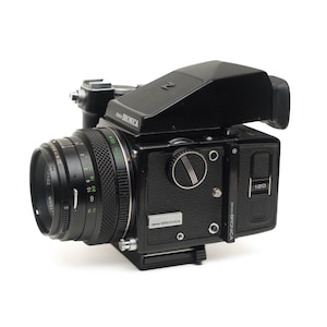 May include: A black medium format film camera with a lens, viewfinder, and a 120 film indicator. The camera has the brand name "Bronica" printed on it. The lens has measurement markings.