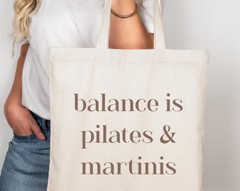 May include: A cream-colored canvas tote bag featuring the brown text "balance is pilates & martinis" printed in a centered serif font. The bag has two long shoulder straps and a rectangular shape, constructed from a woven fabric material.