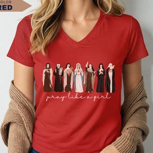 Op de afbeelding: Een rode V-hals T-shirt met een rij ge&iuml;llustreerde figuren en de tekst "pray like a girl" in witte letters. Het shirt heeft korte mouwen en is gemaakt van een zacht materiaal. De kleur van het shirt is rood.
