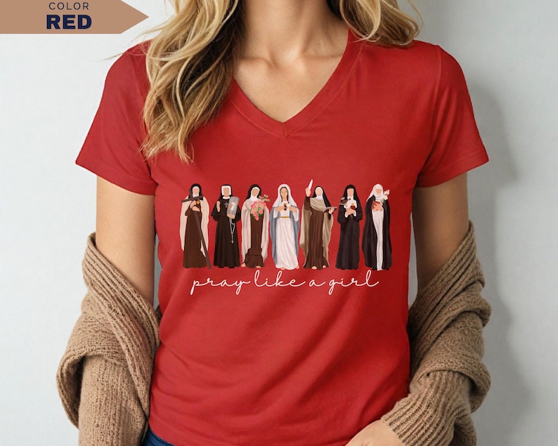 Op de afbeelding: Een rode V-hals T-shirt met een rij ge&iuml;llustreerde figuren en de tekst "pray like a girl" in witte letters. Het shirt heeft korte mouwen en is gemaakt van een zacht materiaal. De kleur van het shirt is rood.