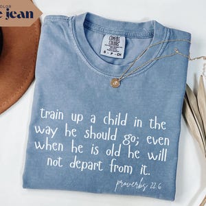 May include: A blue jean colored t-shirt with the text "train up a child in the way he should go; even when he is old he will not depart from it. proverbs 22.6" in white. A gold necklace and a brown hat are also visible.