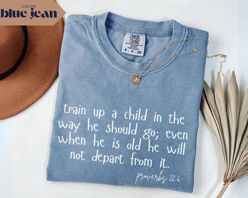 May include: A blue jean colored t-shirt with the text "train up a child in the way he should go; even when he is old he will not depart from it. proverbs 22.6" in white. A gold necklace and a brown hat are also visible.