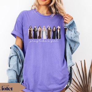 May include: A violet t-shirt featuring a graphic illustration of seven religious figures in traditional habits standing in a row, with the white cursive text "pray like a girl" printed directly underneath the figures on the front of the garment.