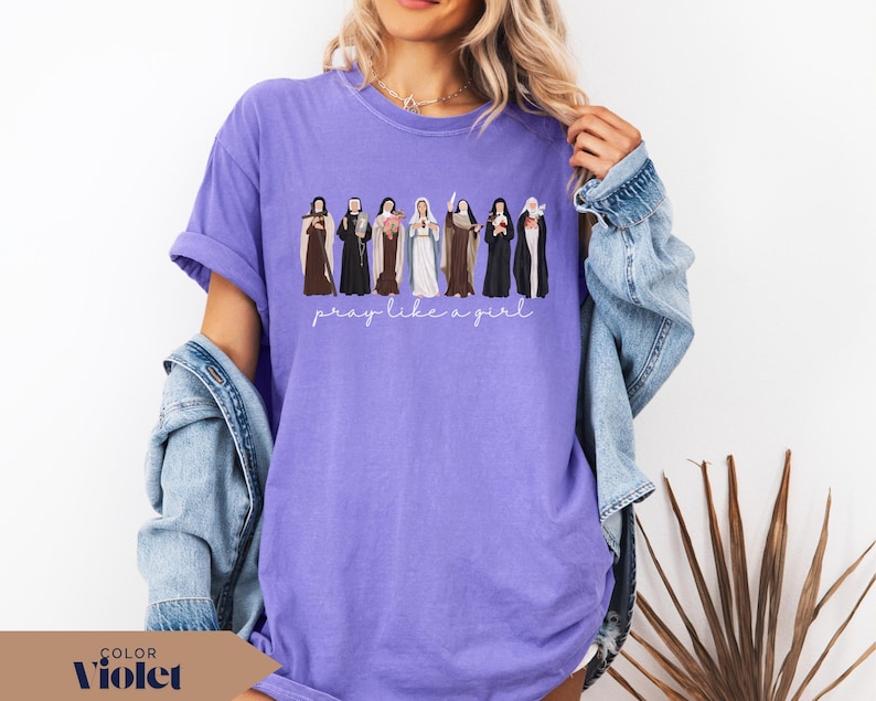 May include: A violet t-shirt featuring a graphic illustration of seven religious figures in traditional habits standing in a row, with the white cursive text "pray like a girl" printed directly underneath the figures on the front of the garment.