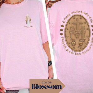 May include: A light pink t-shirt featuring a small illustration of a religious figure on the front. The back of the shirt displays a detailed emblem with text that reads "O Mary conceived without sin, pray for us who have recourse to thee."