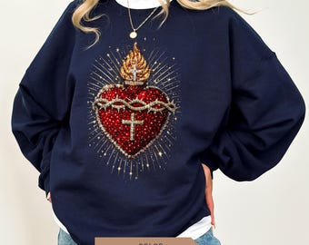 May include: Navy blue sweatshirt featuring a detailed graphic of a Sacred Heart with a crown of thorns, cross, and flames. The design includes glitter-like effects. The text "PRINTED DESIGN - NOT REAL GLITTER" is at the top. The color is Navy.
