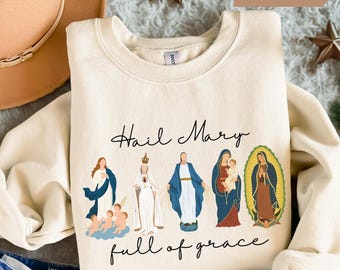 May include: Sand-colored sweatshirt with the text "Hail Mary full of grace" and illustrations of religious figures. The design features figures in blue, white, and red, with a gold halo. The sweatshirt is a crew neck style.