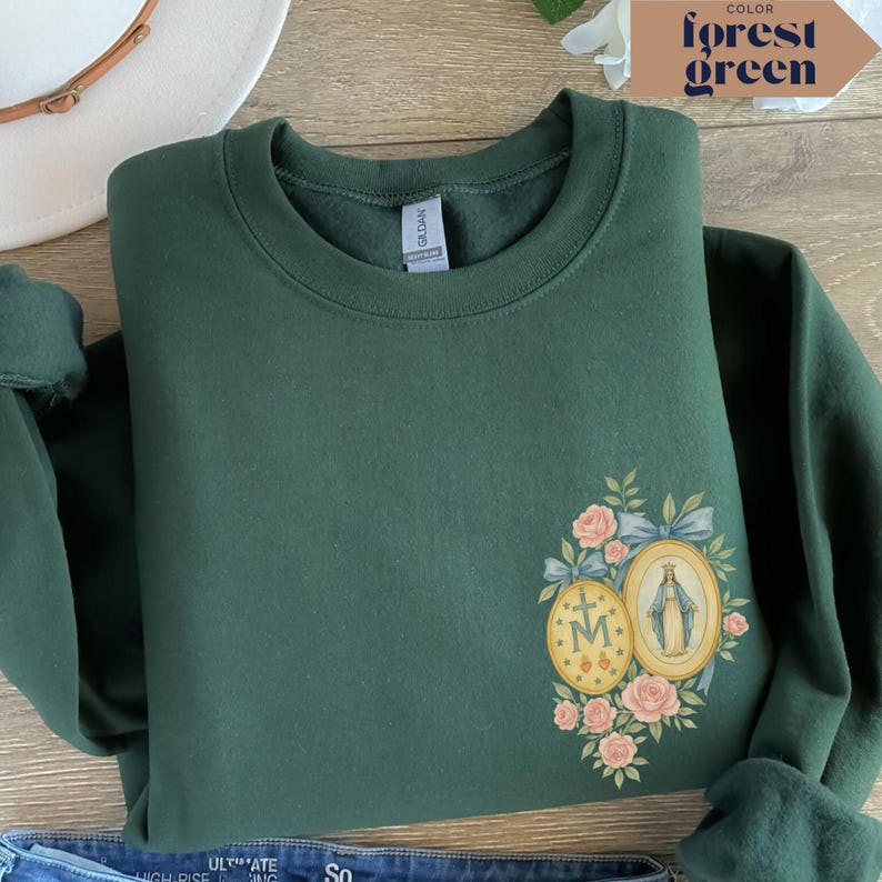 May include: Forest green sweatshirt with a floral and religious medallion design. The design includes pink roses, blue ribbons, and two oval medallions with religious imagery. The sweatshirt is folded, with the sleeves visible.