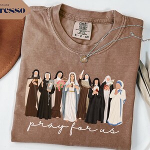 May include: Espresso-colored t-shirt featuring a graphic of several religious figures. The phrase "pray for us" is written in white script across the bottom. A silver necklace with a pendant rests on the shirt.
