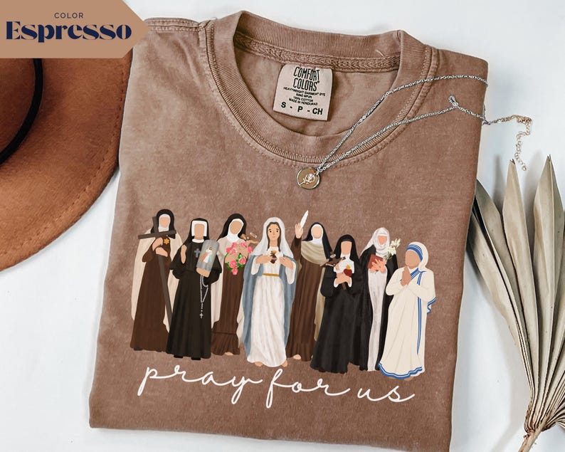 May include: Espresso-colored t-shirt featuring a graphic of several religious figures. The phrase "pray for us" is written in white script across the bottom. A silver necklace with a pendant rests on the shirt.
