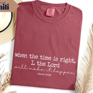 May include: A maroon-colored t-shirt with the text "when the time is right, I, the Lord will make it happen" in white script. The shirt has a crew neck and a tag at the top. A straw hat and decorative plant are in the background.