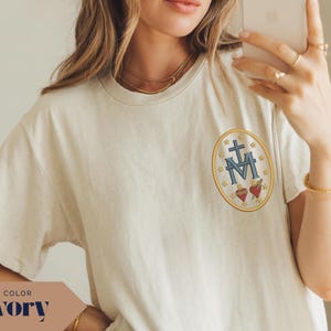 Pu&ograve; includere: T-shirt color avorio con un piccolo stemma ricamato sul petto. Lo stemma presenta una croce, la lettera M e due cuori, il tutto all'interno di un ovale dorato. La maglietta ha maniche corte e girocollo.