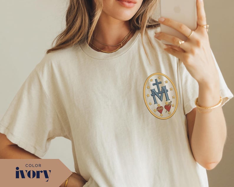 Pu&ograve; includere: T-shirt color avorio con un piccolo stemma ricamato sul petto. Lo stemma presenta una croce, la lettera M e due cuori, il tutto all'interno di un ovale dorato. La maglietta ha maniche corte e girocollo.