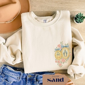 May include: Sand-colored sweatshirt with a floral and religious design. The design includes two oval medallions, one with the letter "M" and the other with a depiction of the Virgin Mary, surrounded by pink roses and blue bows.