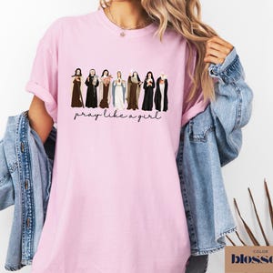 May include: A light pink short-sleeved t-shirt featuring a graphic illustration of eight religious figures in various habits and robes. Below the figures, the text reads "pray like a girl" in a black cursive script font. The shirt has a crew neckline.