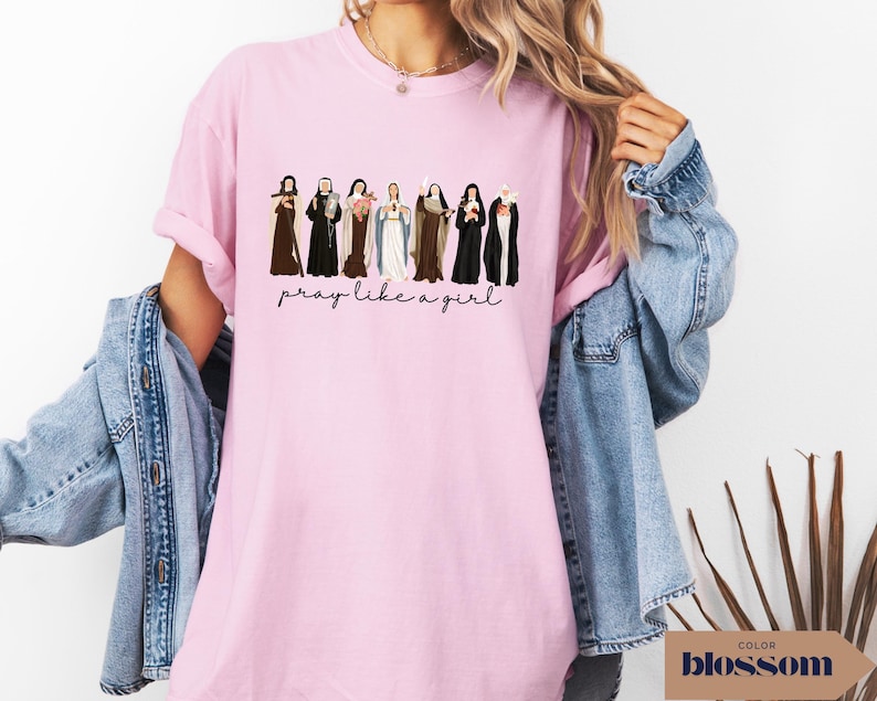 May include: A light pink short-sleeved t-shirt featuring a graphic illustration of eight religious figures in various habits and robes. Below the figures, the text reads "pray like a girl" in a black cursive script font. The shirt has a crew neckline.