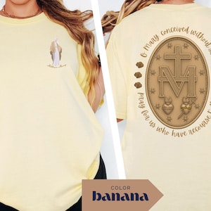 May include: Banana-colored t-shirt with a small illustration on the front and a detailed religious emblem on the back. The emblem features a cross, the letter M, and the text "O Mary conceived without sin, pray for us who have recourse to thee."