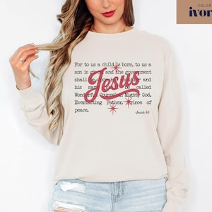 May include: Ivory-colored sweatshirt with the text "For to us a child is born..." and the name "Jesus" in red script. The design includes red starburst accents and the Bible verse Isaiah 9:6.
