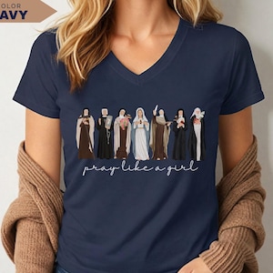 Op de afbeelding: Marineblauwe V-hals T-shirt met een rij ge&iuml;llustreerde religieuze figuren. De figuren zijn gekleed in verschillende gewaden. De zin "pray like a girl" is in cursief geschreven. Het T-shirt is effen marineblauw.