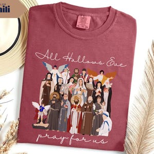 May include: A maroon t-shirt with the words "All Hallows Eve pray for us" in white script. The shirt features a graphic of various religious figures. A straw hat and dried plants are in the background.