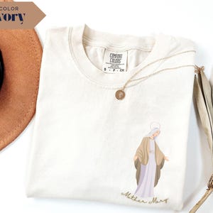 May include: Ivory-colored Comfort Colors t-shirt featuring an illustration of Mother Mary in a light brown and lavender color scheme. The words "Mother Mary" are written in a cursive font below the illustration. A gold necklace with a pendant is draped across the shirt.