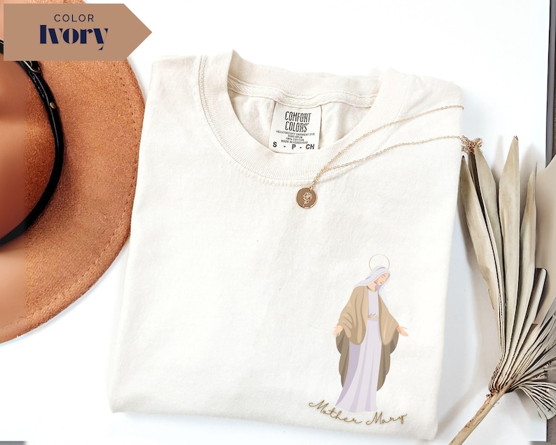 May include: Ivory-colored Comfort Colors t-shirt featuring an illustration of Mother Mary in a light brown and lavender color scheme. The words "Mother Mary" are written in a cursive font below the illustration. A gold necklace with a pendant is draped across the shirt.
