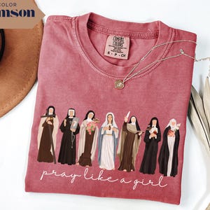May include: A crimson short-sleeve t-shirt featuring a graphic of seven religious figures in various habits and robes. Below the figures, the text reads "pray like a girl" in white cursive script. The shirt has a Comfort Colors brand label.