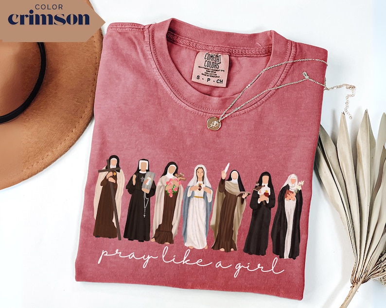 May include: A crimson short-sleeve t-shirt featuring a graphic of seven religious figures in various habits and robes. Below the figures, the text reads "pray like a girl" in white cursive script. The shirt has a Comfort Colors brand label.