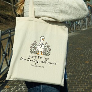May include: Cream-colored tote bag featuring a cartoon duck with folded hands, surrounded by daisies. The text "sorry I'm late, His timing, not mine, Ecclesiastes 3:1" is printed below the duck. The bag is held by a person.