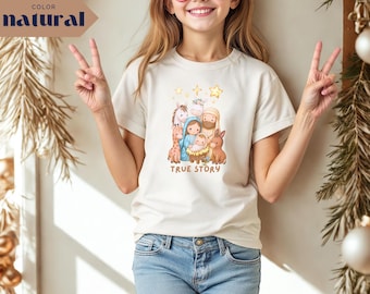 May include: A cream-colored t-shirt featuring a colorful illustration of a nativity scene with the text "TRUE STORY" in a playful font. The design includes figures of Mary, Joseph, baby Jesus, animals, and stars. The shirt is worn by a person.