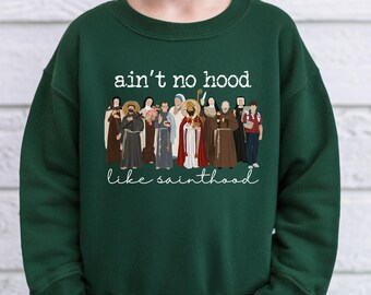 May include: Forest green sweatshirt with the text "ain't no hood like sainthood" in white script. The graphic features illustrations of various religious figures. The color of the sweatshirt is labeled "forest green" in a tan banner.