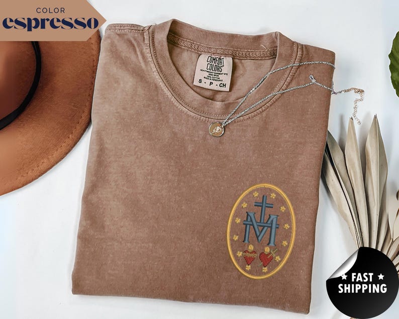 Pu&ograve; includere: T-shirt color espresso con un design ovale ricamato con una croce, le lettere "M" e "V" e due cuori. Una collana d'argento con un ciondolo poggia sulla maglietta. La maglietta &egrave; piegata e un cappello marrone &egrave; parzialmente visibile.