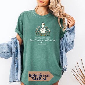 Op de afbeelding: Lichtgroen T-shirt met een afbeelding van een biddende eend omringd door madeliefjes. De tekst luidt "sorry I'm late, His timing, not mine, Ecclesiastes 3:1". Het shirt is maat XL.
