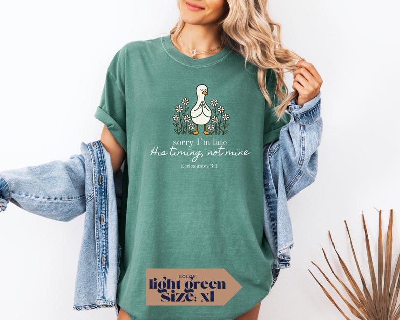 Op de afbeelding: Lichtgroen T-shirt met een afbeelding van een biddende eend omringd door madeliefjes. De tekst luidt "sorry I'm late, His timing, not mine, Ecclesiastes 3:1". Het shirt is maat XL.